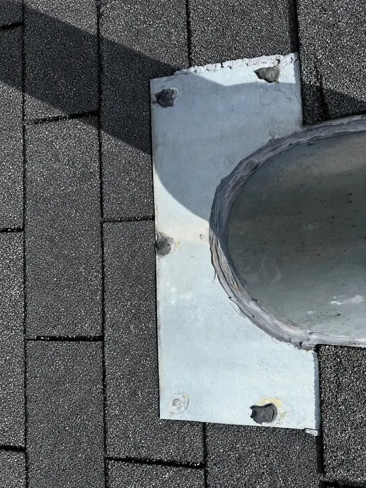 Metal flashing plate around a gas vent pipe on a shingle roof in Holly Springs, North Carolina, with exposed nail heads, before the leak repair